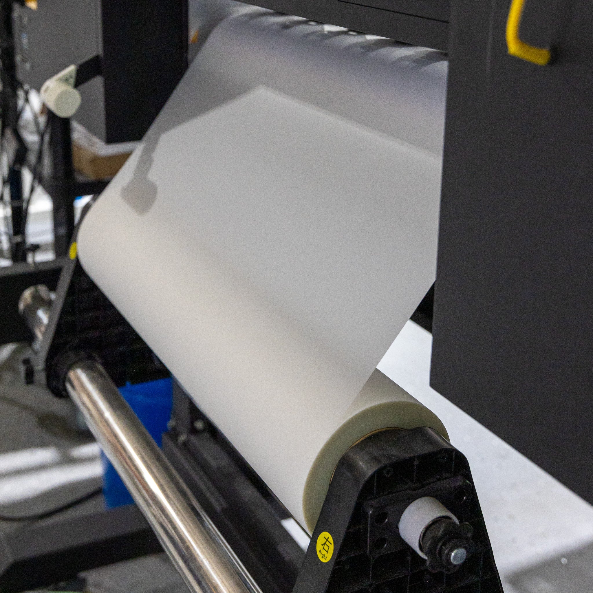 A 24in Hot Peel DTF Film - Double Side Matte by DTF Locker feeds through a large-format printer's rollers, with black components visible in an industrial or office setting.